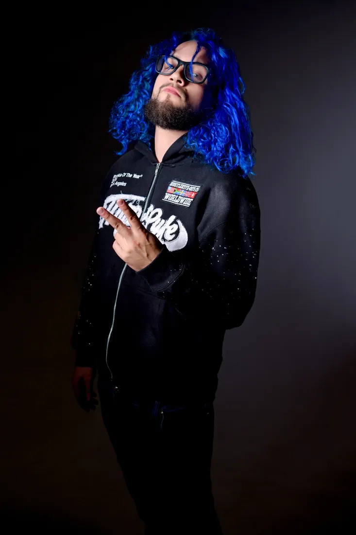 Fly South in black with bright blue hair and glasses, one hand raised, against a dark studio backdrop